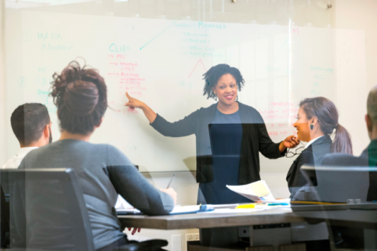 An image of a person presenting information on a whiteboard to a group of people.