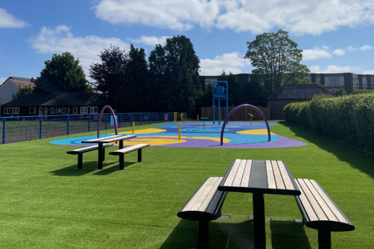 Churchfield splash pad with two picnic tables, green artificial grass and the water play area