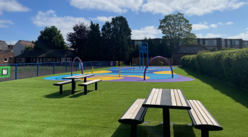 Churchfield splash pad with two picnic tables, green artificial grass and the water play area